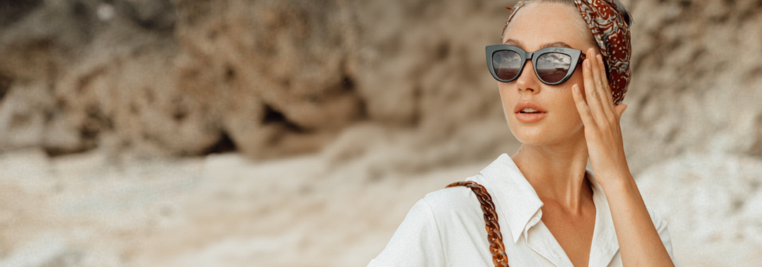 Woman wearing a white summer dress and sunglasses against a rocky coastal background.